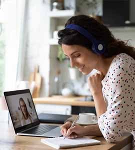 Woman in home office seated at laptop video call with notepad and pen